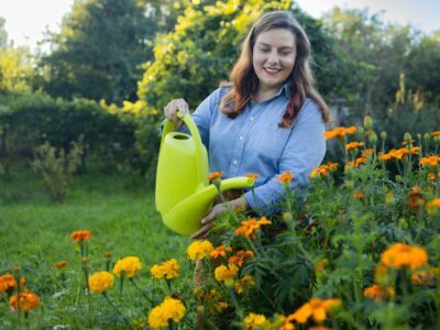 Cheerful attractive young woman gardener pouring flowers with green watering can in garden