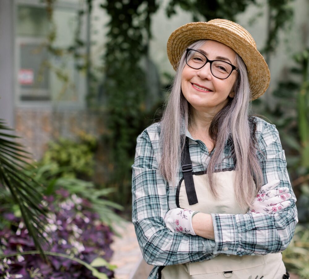 Close up of happy smiling senior lady gardener with long grey hair, wearing straw hat